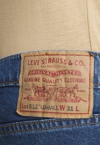 Denimová etiketa Levi Strauss & Co. má červený text na béžovém papíru a obsahuje detaily "Originální nýtované", "Kvalitní oděvy", Lot 512, W 31 L.