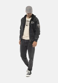 Veste à capuche zippée en polaire gris foncé avec une texture lisse, ornée de patches sur les manches ; associée à un jean gris et des baskets noires.