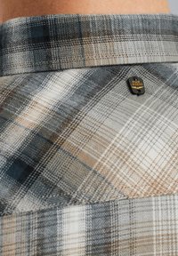 Camisa a cuadros con una mezcla de tejido suave, con cuello, en tonos neutros de gris, azul y beige, y una pequeña etiqueta negra de la marca.