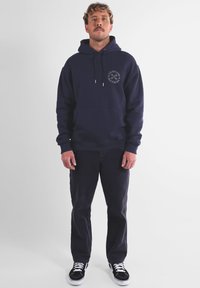 Hoodie marine avec poche avant, capuche à cordon et design en logo circulaire. Associé à un pantalon sombre et des baskets noires. Look propre et décontracté.