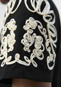 Svart tygskjorta med intrikata off-white broderade detaljer, med virvlande och loopade mönster längs ärmarna.