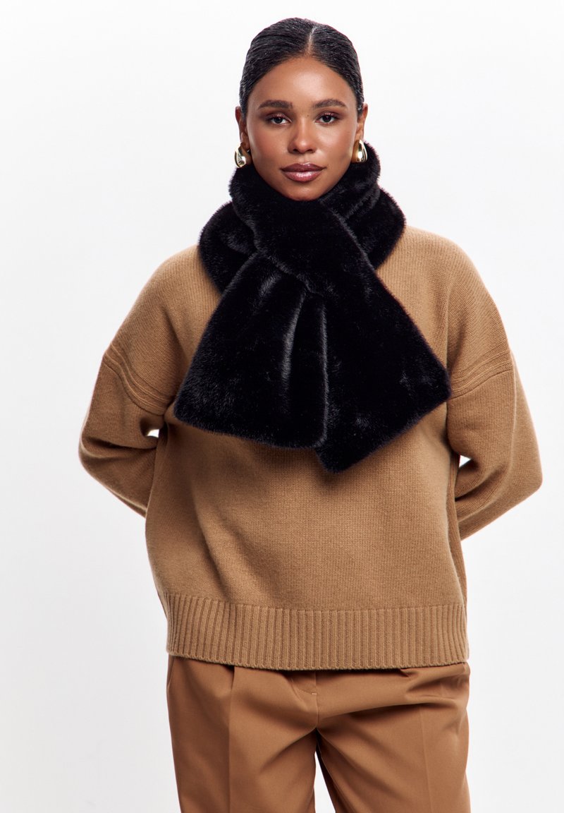 Sort faux fur tørklæde med en blød tekstur, arrangeret i en løkke, båret over en tan strikket sweater, komplementeret af brune bukser.