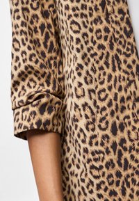 Le tissu à imprimé léopard présente des taches brun foncé sur un fond beige ; la manche est retroussée, dévoilant une texture douce et une finition lisse.