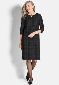 Schwarzes knielanges Kleid aus strukturiertem Stoff mit subtilen Karomuster, Rundhalsausschnitt und dreiviertellangen Ärmeln, kombiniert mit schwarzen High Heels.