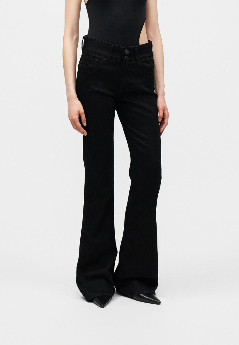 Άτομο που φοράει ψηλόμεσα μαύρα καμπάνα παντελόνια με κούμπωμα με κουμπιά και μαύρα παπούτσια με μυτερή μύτη σε απλό φόντο.