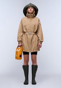 Veste imperméable beiger avec capuche verte, taille cintrée et cordons ajustables. Associée à un short noir, des bottes vertes et un sac orange.