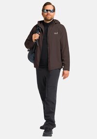 Veste zippée marron sur un haut noir à manches longues, associée à un pantalon et des chaussures noires ; portant des lunettes de soleil sombres, tenant un sac bleu.