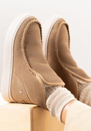 Baskets slip-on en daim beige avec une doublure en polaire douce, présentant un détail cousu le long des bords et une semelle en caoutchouc blanche.