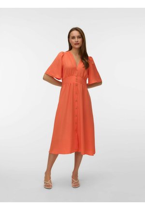 Žena stojaca s rukami za chrbtom, oblečená v koralových midi šatách s gombíkmi vpredu, krátkymi rukávmi a otvorenými opätkami.