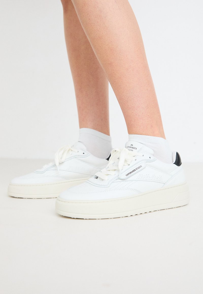 Weiße Leder-Sneaker mit einer robusten Sohle, durchbrochenen Details, schwarzen Fersenelementen und weißen Schnürsenkeln. Getragen mit grauen Crew-Socken.