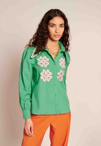Zeleni bombažni srajca z roza vezenimi cvetličnimi vzorci, koničast ovratnik in zapenjanje spredaj; v kombinaciji z oranžnimi hlačami. Teksturna tkanina.