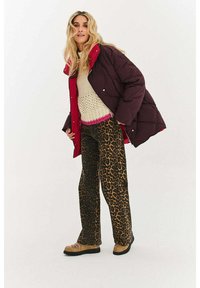 Pikowana bordowa kurtka z czerwonym kołnierzem na beżowym swetrze z różowym obszyciem, zestawiona z szerokimi spodniami w panterkę i brązowymi butami.