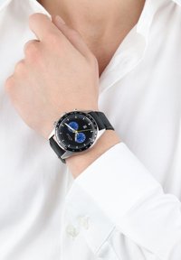 Schwarze Chronographuhr mit blauen und gelben Akzenten, silbernem Gehäuse und Gummiband, getragen an einer Hand in der Nähe eines weißen Hemdes.