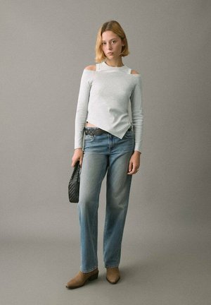 Top gri deschis cu mâneci lungi cu decupaje la umeri, asortat cu blugi evazați de spălare medie și cizme maro până la gleznă. Ține o geantă neagră împletită.