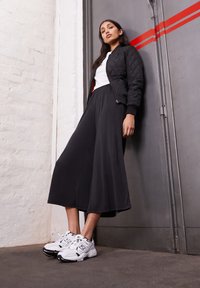 Young woman leaning against gray door with red stripes wearing black quilted jacket, wide-leg black pants, white shirt, and white sneakers.