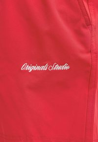 Tissu rouge avec texte brodé en blanc "Originals Studio" et couture visible ainsi que détails de poche sur le côté droit.