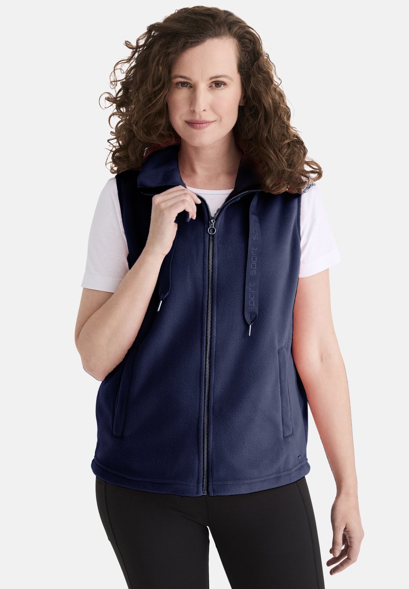 Marineblauer Fleece-Weste mit durchgehendem Reißverschluss, Stehkragen und seitlichen Taschen. Mit Zipper-Details und logobezogenen Akzenten. Getragen über einem weißen Hemd.
