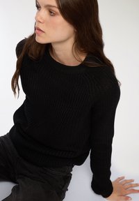 Maglione nero lavorato a maglia con texture a coste, scollatura rotonda e maniche lunghe. Indossato con pantaloni scuri. Modello seduto su uno sfondo chiaro.