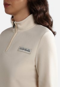Sudadera de forro polar en color beige claro, con un cuello de media cremallera y un parche rectangular de logo en el pecho. Textura suave con un acabado liso.