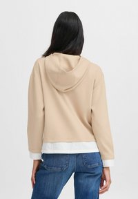 Beige cropped hoodie met een trekkoordcapuchon, met een zachte textuur en een contrasterende witte zoom. Gedragen over blauwe spijkerbroek.