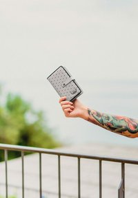 Šedá peněženka s černými puntíky, skládací design, zabezpečená páskem se zacvakávacím uzávěrem a s logem jako akcentem.