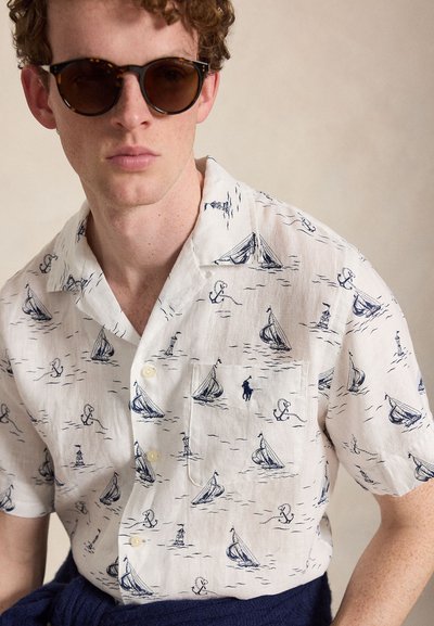 Jeune homme portant des lunettes de soleil écaille et une chemise blanche à manches courtes avec des motifs de voilier, ancre et phare bleu marine.
