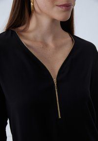 Black top featuring a V-neck and a gold zipper detail. Smooth fabric with a close fit, highlighting a minimalist design without patterns.