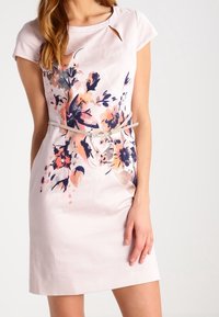 Vaaleanpunainen mekko lyhyillä hihoilla, kukkakuosi vaaleanpunaisissa ja sinisissä sävyissä, jossa on avainreikäyksityiskohta kaula-aukossa ja vyö vyötäröllä.