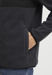 Chaqueta de forro polar gris oscuro con cierre frontal, bolsillos laterales y detalles en negro en los hombros y los puños. Tejido suave y texturizado.