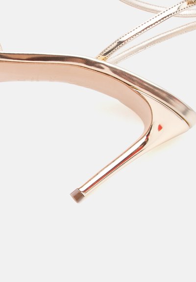 Χρυσό μεταλλικό στιλέτο με λεπτά λουράκια και καφέ άκρη σε λευκό φόντο, με εμφανή μέρος της σόλας του παπουτσιού.