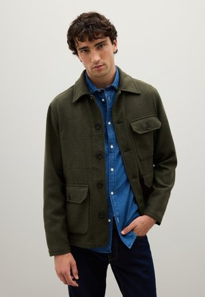 Young man wearing a green buttoned jacket over a blue denim shirt and dark jeans, standing against a plain light background.