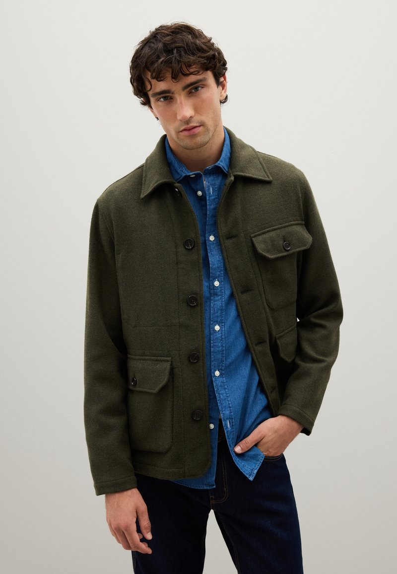 Young man wearing a green buttoned jacket over a blue denim shirt and dark jeans, standing against a plain light background.
