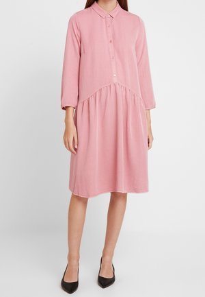 Femme portant une robe rose ample jusqu'aux genoux avec des manches longues, un col, des boutons sur le devant et des talons noirs pointus, debout devant un fond uni.