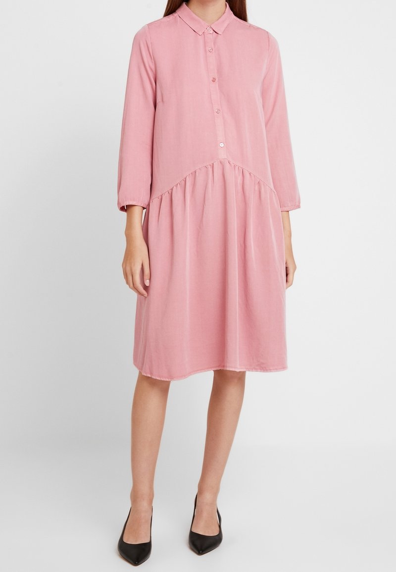 Femme portant une robe rose ample jusqu'aux genoux avec des manches longues, un col, des boutons sur le devant et des talons noirs pointus, debout devant un fond uni.