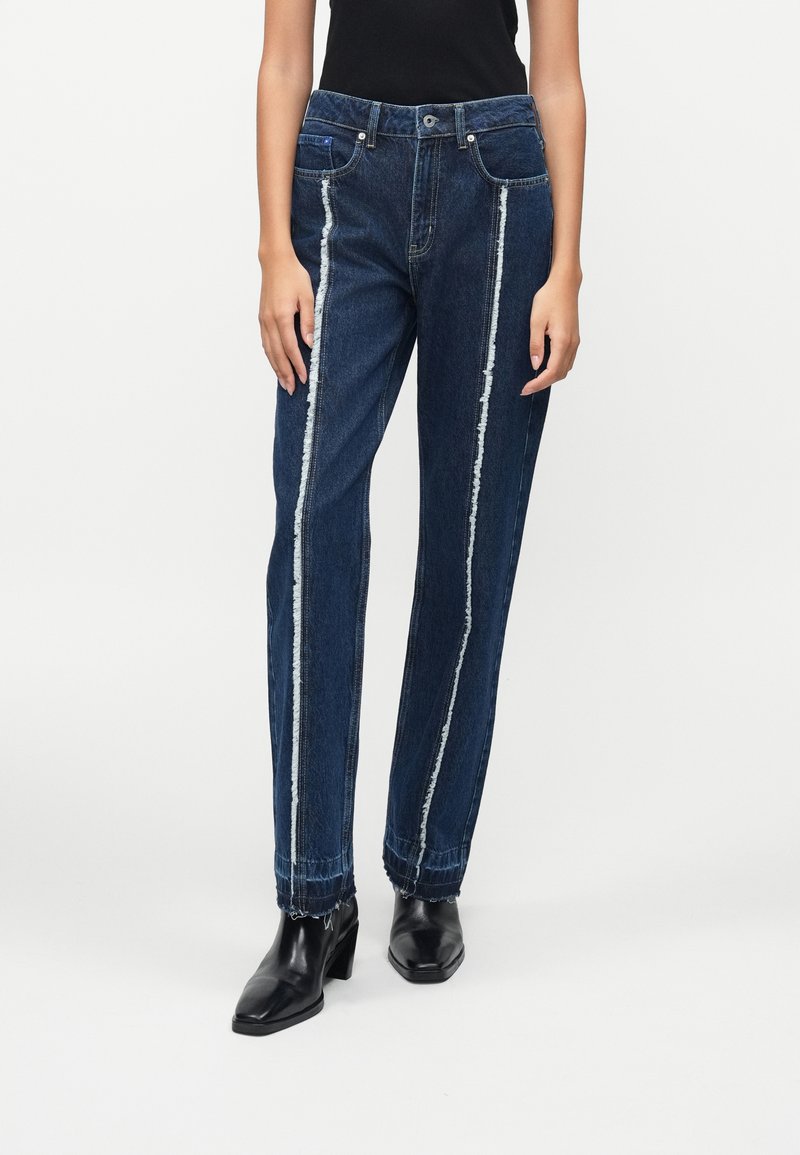 Jeans de denim azul escuro com riscas laterais brancas desgastadas, ajuste solto e punhos afunilados, combinados com botas pretas de salto agudo.