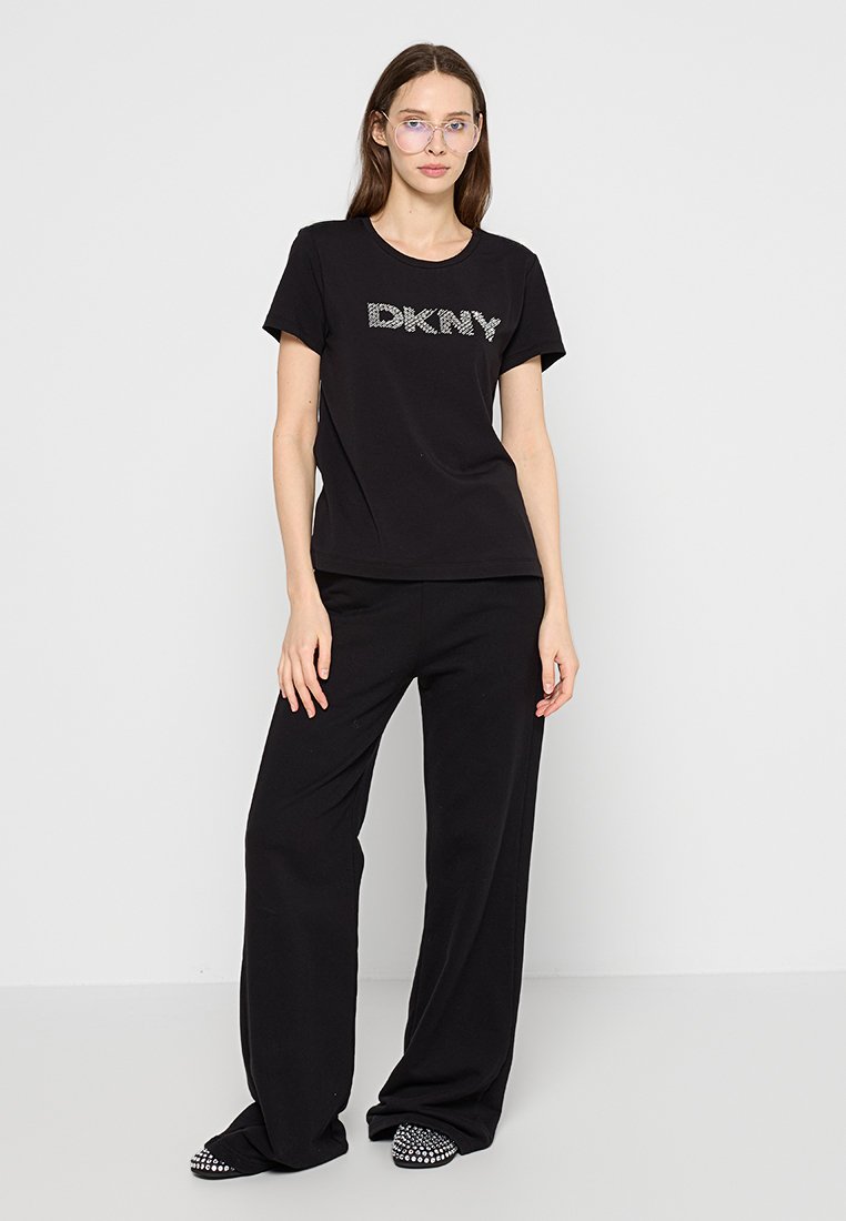 Zwarte t-shirt met glanzend DKNY-logo, gecombineerd met zwarte wijde broek en decoratieve zwarte instappers met studs.