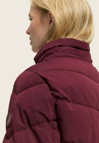 Bordo puf jakna s visokim ovratnikom, s ušivenim dijelovima i kružnom logom na lijevom rukavu. Glatka, mat tekstura.