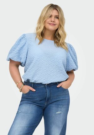 Femme aux cheveux blonds portant un haut bleu clair texturé à manches bouffantes et un jean bleu, debout les mains dans les poches devant un fond uni.