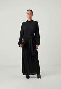 Črna dolga obleka z dolgimi rokavi, visokim ovratnikom in zavezanem pasu. Gladka tkanina, dolžina do tal, obuta z majhno torbico in čevlji s peto.