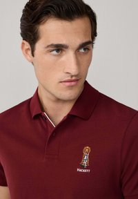 Burgundské polo tričko s textúrovaným povrchom, obsahuje podrobný vyšívaný logotip psa a text 'Hackett' na hrudi, klasický golier s gombíkmi.