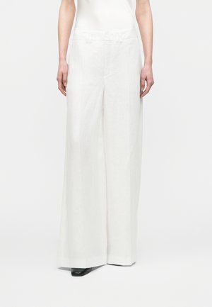 Persona vistiendo pantalones blancos de talle alto y pierna ancha con zapatos negros puntiagudos, de pie contra un fondo blanco liso.