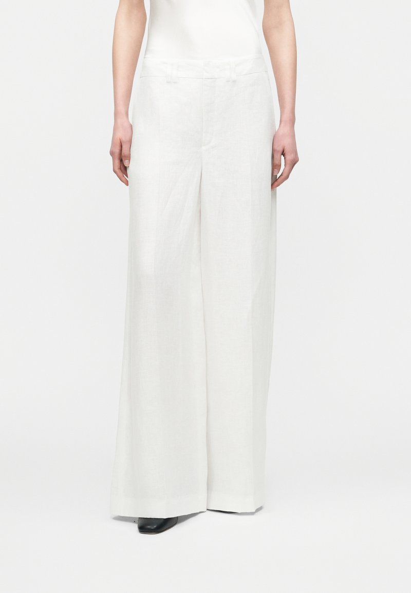 Personne portant un pantalon blanc taille haute à jambes larges avec des chaussures noires à bouts pointus, debout devant un fond blanc uni.