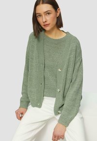 Cardigan en tricot vert avec un col en V et une fermeture à boutons, assorti à un pull. Le tissu présente un motif texturé et une finition douce.