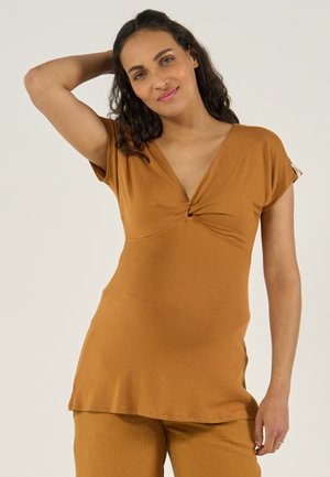 Vrouw met krullend donker haar, gekleed in een roestkleurige tuniek met V-hals en broek, die één arm achter haar hoofd legt en zachtjes glimlacht.