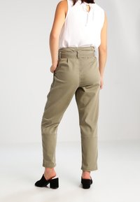 Kvinna i högmidjade olivgröna byxor med bälte och vit ärmlös blus, står med händerna i fickorna och vänder sig bort.