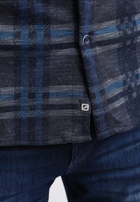 Gros plan sur une chemise à carreaux bleu foncé et gris boutonnée sur un jean en denim bleu avec une petite étiquette de marque noire près de l'ourlet.