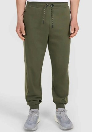 Mężczyzna noszący oliwkowe spodnie dresowe ze sznurkiem w pasie i boczną kieszenią na zamek, w połączeniu z jasnoszarymi butami sportowymi.