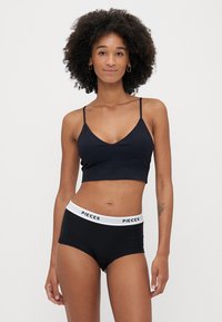 Fekete bralette vékony pántokkal és V nyakkivágással, fekete hipster rövidnadrággal párosítva, amin egy fehér derékszíj található "PIECES" logóval.