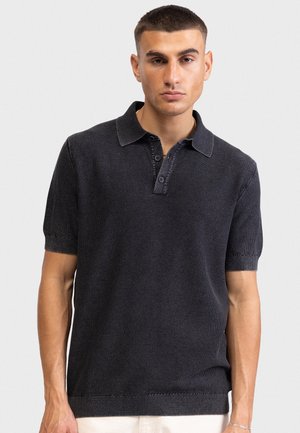 Νεαρός άνδρας με κοντά μαλλιά, φορώντας σκούρο πλεκτό πόλο με κοντά μανίκια, στέκεται μπροστά σε απλό ανοιχτόχρωμο φόντο.