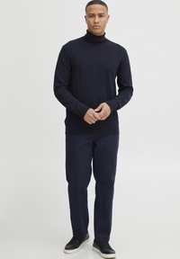 Navy coltrui van breistof, gecombineerd met donkerblauwe broek. Zwarte sneakers met witte zolen complementeren de outfit.